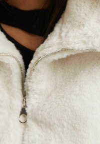 Vit fleece zip-up jacka med en mjuk textur och silverdetaljer på dragkedjan. Närbild som fokuserar på den övre delen och kragen.