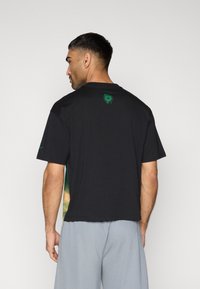 Μαύρο βαμβακερό μπλουζάκι με πράσινο λογότυπο στην πλάτη και πολύχρωμο γραφικό στα πλαϊνά. Κοντά μανίκια και χαλαρή εφαρμογή.