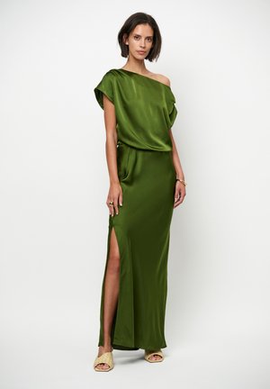 Groene satijnen jurk met één off-shoulder mouw, een zijsplit en een losse pasvorm. Model draagt neutrale slip-on sandalen en een minimalistische ring.