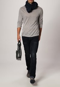 Hombre caminando con una camisa de manga larga gris claro, jeans oscuros, zapatos grises, una bufanda negra y llevando un maletín de cuero negro.