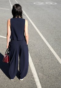 Námořnický overal s přiléhavým topem bez rukávů a širokými nohavicemi. Hladká tkanina, minimalistický design. Drží malou červenou kabelku.