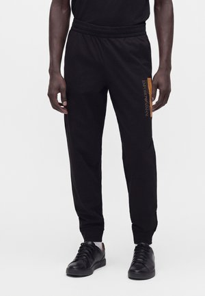 Homme portant un pantalon de survêtement noir avec un logo Emporio Armani orange sur la cuisse et des baskets en cuir noir.