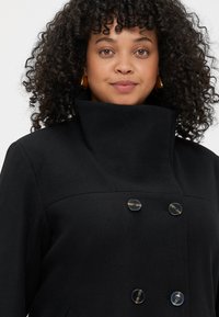 Cappotto in misto lana nero con colletto alto, chiusura con bottoni doppi e bottoni rotondi. Il tessuto testurizzato presenta linee pulite.