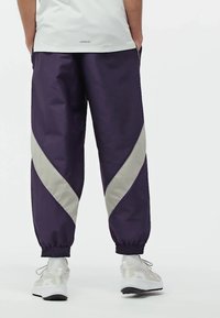 Fialové joggingové nohavice s ľahkým šedým diagonálnym pruhom, elastickými manžetami a hladkým povrchom. Nosia sa s bielymi teniskami.