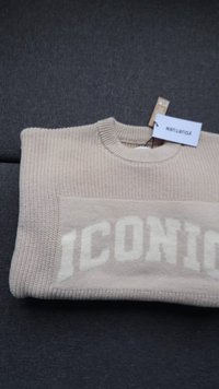 Beige kötött pulóver összehajtva sötét szövet felületén, az "ICONIC" felirattal és két ruhacímkével a nyak körüli területen.