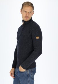 Navy ribbed sweater with a high collar and quarter zipper, features a textured knit and a small brown logo patch on the left sleeve.