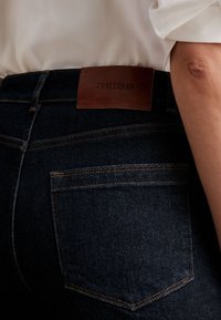 Tmavomodré rifle z denimu s hnedou koženou nášivkou označenou "REISS" na páse, nosené s bielou košeľou a ukazujúce časť ohnutého ramena.