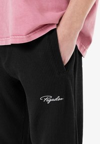 Sorte ribbede bukser med sidelommer og hvid "Pegador" broderi på venstre lår, parret med en lyserød T-shirt med korte ærmer.