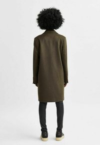 Cappotto verde scuro in misto lana con taglio dritto, maniche lunghe e dettagli con bottoni, abbinato a pantaloni neri e scarpe scure.