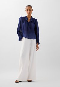 Bluză eficientă de culoare bleu marin, cu guler volan, detalii cu panglici la mâneci, asortată cu pantaloni albi cu picioare largi și sandale negre cu talpă plată.