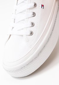 Detailní záběr na bílé plátěné tenisky s bílými tkaničkami, stříbrnými oky, texturovanou silnou podrážkou a malým vyšitým červeným a modrým logem.