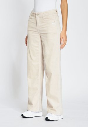 Personne portant un pantalon en velours côtelé beige à jambes larges et des baskets blanches, debout devant un fond clair uni.