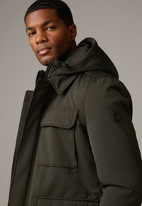 Olivgrüne gepolsterte Jacke mit großer Kapuze, zwei Fronttaschen und einem schwarzen Logo auf dem linken Ärmel. Glatte Textur und taillierte Passform.