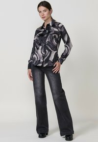 Camicia a maniche lunghe a bottoni nera e grigia a fantasia, abbinata a jeans a zampa d'elefante scuri, indossata con stivali neri con tacco.
