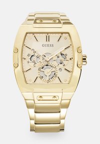 Gullfarget Guess-klokke med rektangulært klokkeansikt, beige urskive, kronograf underdials og en kraftig metallarmbånd. Har markante timemarkører.