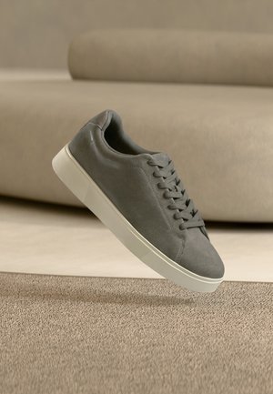 Baskets en daim gris avec semelle blanche et lacets flottant au-dessus d'un tapis beige texturé dans une pièce aux tons neutres.