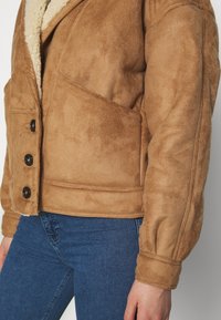 Brun fuskmockajacka med fårskinnskrage, med fyra svarta knappar, sidofickor och en något kortare design. Strukturerad yta.