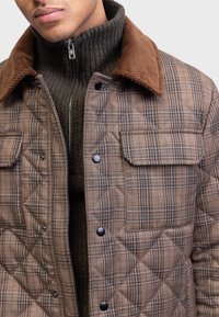 Homme portant une veste matelassée à carreaux marron avec un col en velours côtelé par-dessus un pull zippé côtelé vert foncé, montré du menton à la taille.