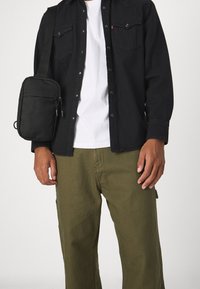 Black button-up shirt over a white t-shirt, olive green pants, and a small black crossbody bag with a zipper detail.