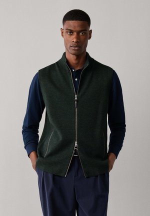 Hombre con un chaleco verde oscuro sin mangas con cremallera sobre una camisa azul marino de manga larga, con las manos en los bolsillos del chaleco, sobre un fondo liso.