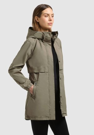 Nainen pukeutuneena oliivinvihreään hupulliseen sadetakkiin, jossa vetoketju ja nepparit, kädet taskuissa, seisoo yksiväristä vaaleanharmaata taustaa vasten.