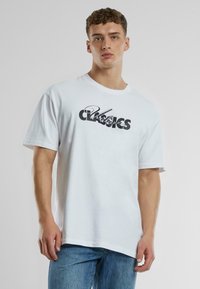 Balta medvilninė marškinėliai su apvalia iškirpte, su juodu grafikiniu užrašu "Urban Classics" per krūtinę. Trumpomis rankovėmis, laisvas kirpimas.