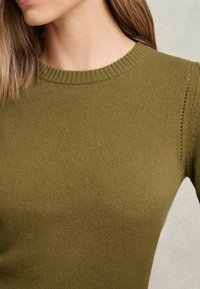 Detailní záběr na ženu v zeleném svetru s žebrovaným výstřihem, na kterém je vidět část jejího krku a ramene na jednobarevném pozadí.