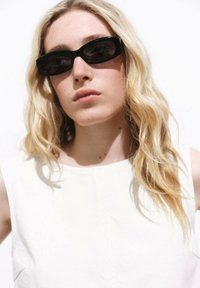 Schwarze, rechteckige Sonnenbrille mit flachem Oberteil. Sie haben dunkle Linsen und eine matte Oberfläche. Der Hintergrund ist reinweiß.