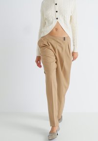 Pantalons beige ajustés avec une taille haute, des plis latéraux et deux boutons noirs, associés à un cardigan crème en tricot torsadé.