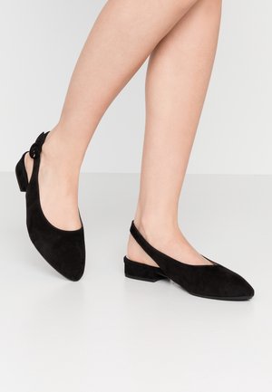 Nogi w czarnych zamszowych butach typu slingback na niskim obcasie, z ostrymi noskami i ozdobną klamrą z boku.
