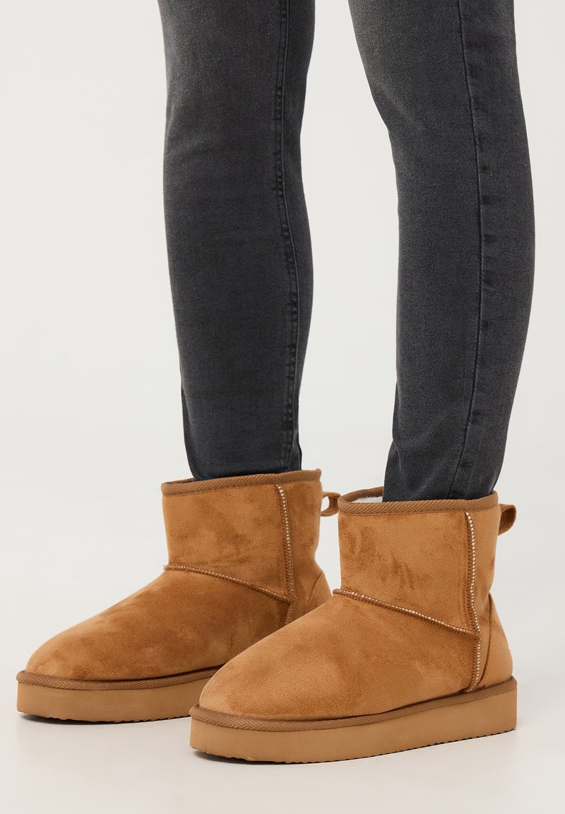 Stivali ankle in suede beige con punta rotonda e suola piatta; presentano un dettaglio cucito e un'ansa sul retro per facilitarne l'indosso.