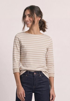 Femme souriante regardant sur le côté, portant une chemise à manches longues beige et blanche rayée et un jean bleu foncé, les cheveux attachés en arrière.