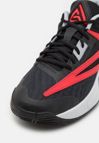 Chaussure de sport noire avec des accents rouges et blancs, empeigne en mesh détaillée, lacets noirs et semelle noire texturée sur fond blanc.