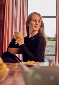 Black long-sleeve top with small metallic accent; model holding a white mug, seated near a table with lemons and pastries. Red striped curtains in background.