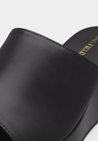 Μαύρο σανδάλι με λείο, φαρδύ δερμάτινο λουράκι και επίπεδη σόλα, με χρυσό κείμενο του εμπορικού σήματος στο πέλμα.