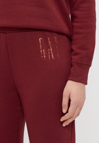 Burgundské tepláky s hladkou texturou, s lesklým růžově zlatým logem "GAP" na levém boku a volným střihem.