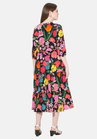 Kwiatowa sukienka maxi z rękawami o długości trzech czwarte, czarne tło, kolorowe kwiaty, warstwowa spódnica i taliowany pas. Białe sandały na obcasie.