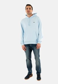 Sudadera azul clara con un bolsillo frontal, que presenta un pequeño logo verde. Combinada con jeans oscuros y zapatillas negras con acentos marrones.