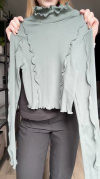 Green ribbed long-sleeve top with a high ruffled collar, featuring wavy detailing along the sleeves and a cropped hem.