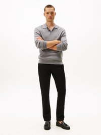 Suéter cinza de malha com colarinho, calças pretas e loafers pretos. Modelo está de braços cruzados contra um fundo branco.