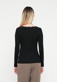 Top negro de manga larga con forma ajustada y un escote de encaje con borde en forma de ola, combinado con pantalones beige. Textura suave.