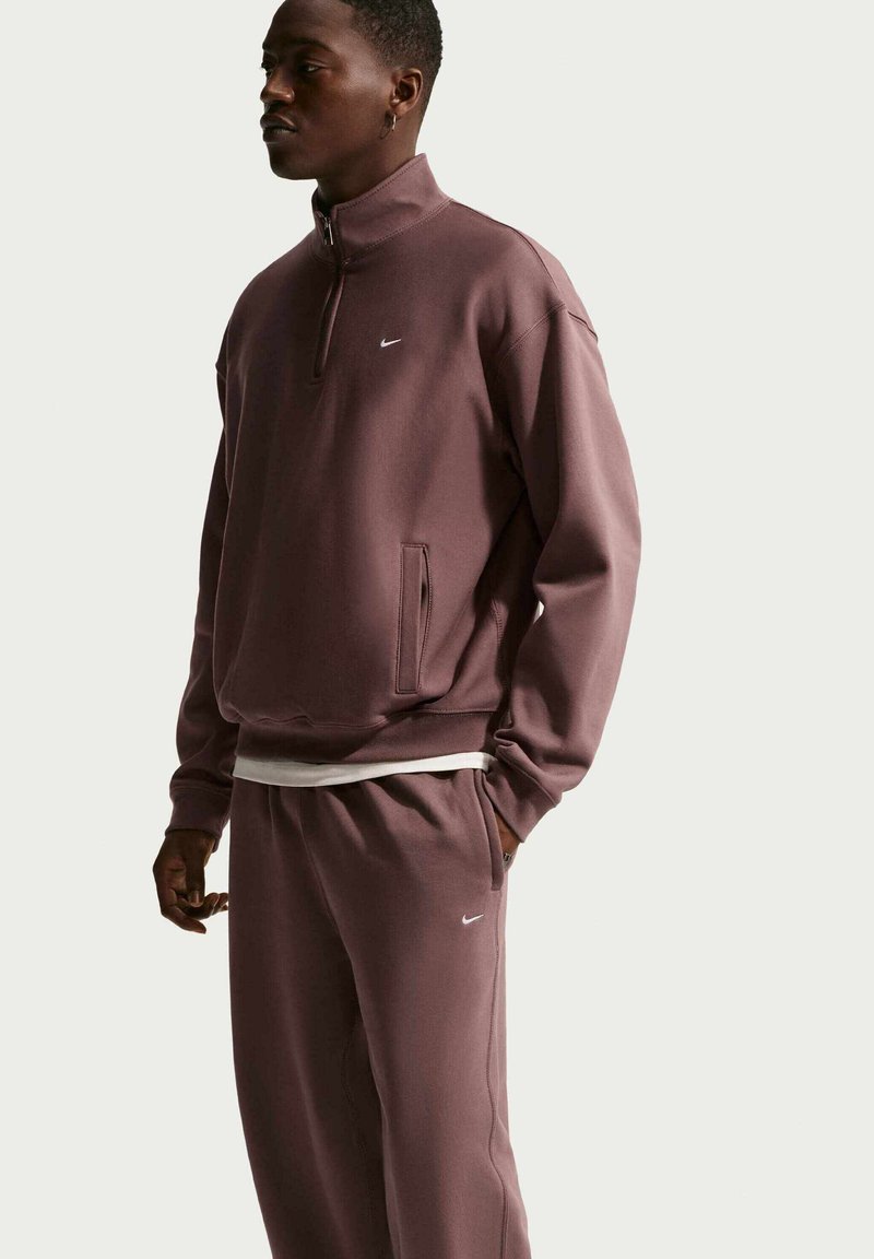 Man met een mauve Nike-trainingspak, een sweater met ritssluiting bij de kraag en bijpassende broek, staand tegen een effen lichte achtergrond.