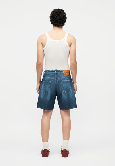 Άνδρας με σγουρά μαλλιά στέκεται στραμμένος με την πλάτη, φοράει λευκή αμάνικη μπλούζα, τζιν σορτς μέχρι το γόνατο, λευκές κάλτσες και κόκκινα αθλητικά παπούτσια σε απλό φόντο.