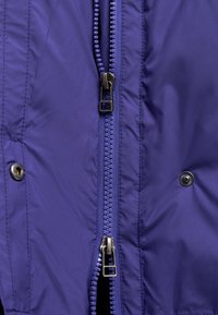 Tkanina ljubičaste izolirane jakne s centralnim patentnim zatvaračem i kopčanjem na dugmad. Patentni zatvarač ima metalnu vuču, a dodaci uključuju prošivene detalje.