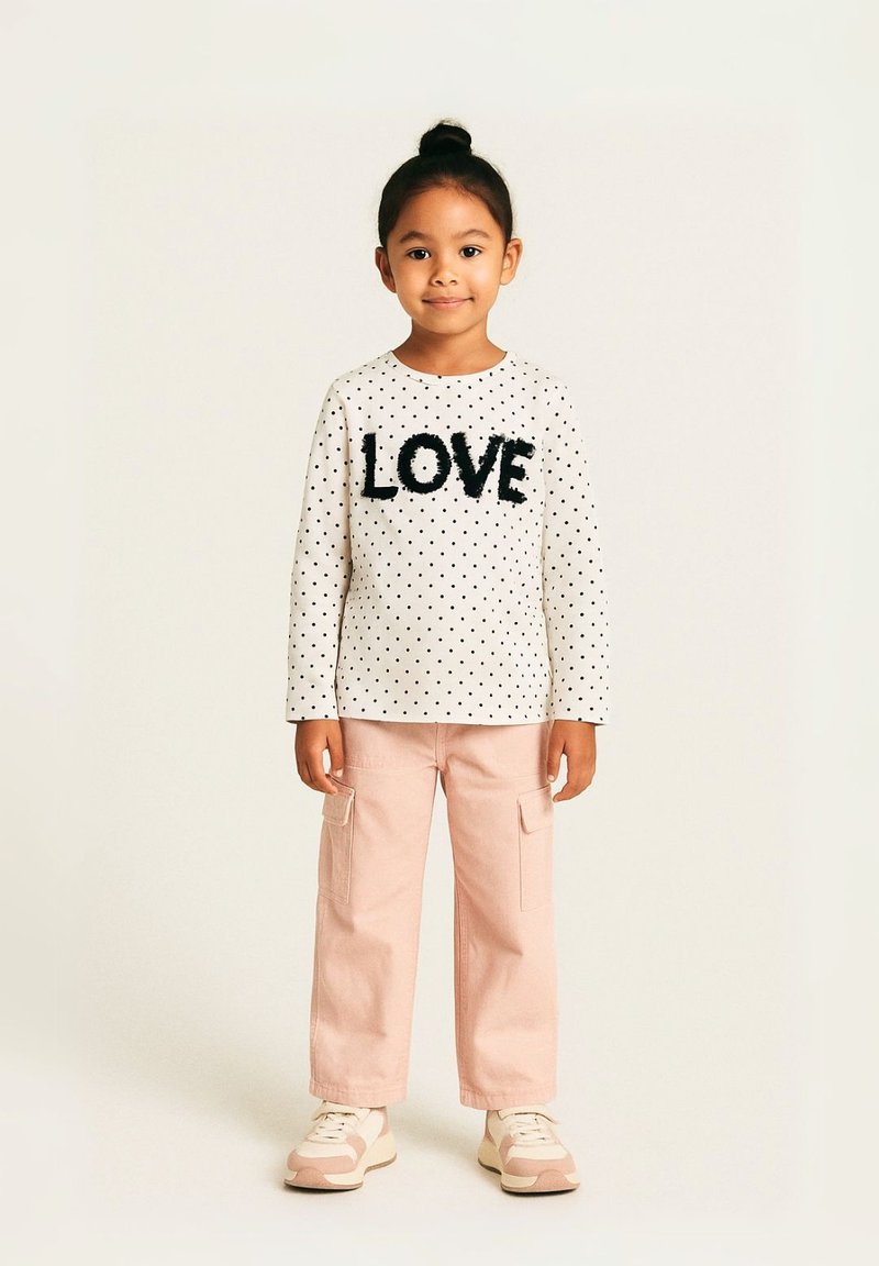 Haut à manches longues blanc avec des pois noirs et un lettrage texturé "LOVE", associé à un pantalon cargo rose et des baskets claires.