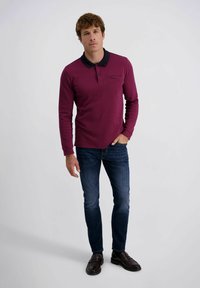 Burgunderrotes Langarm-Polohemd mit navyfarbenem Kragen, Detail mit Brusttasche, kombiniert mit schmal geschnittenen blauen Jeans und dunkelbraunen Loafers.