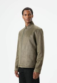 Olivgrüne Jacke aus Kunstwildleder mit Stehkragen, Frontreißverschluss und Seitentaschen. Glatte Textur und taillierter Schnitt.