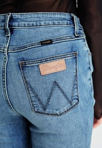 Μπλε τζιν παντελόνι με δερμάτινη ετικέτα με την ένδειξη "Wrangler" και χαρακτηριστικό σχέδιο πορτοφολιού με διπλή ραφή σε σχήμα 'W'.