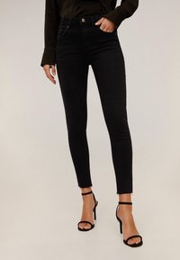 Mujer vestida con jeans skinny de tiro alto negros con dobladillos deshilachados y sandalias de tacón alto negras con tiras, de pie contra un fondo neutro.