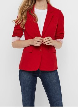 Kvinde iført rød blazer over hvid skjorte med opkørte ærmer og mørkeblå jeans, stående mod ensfarvet baggrund.
